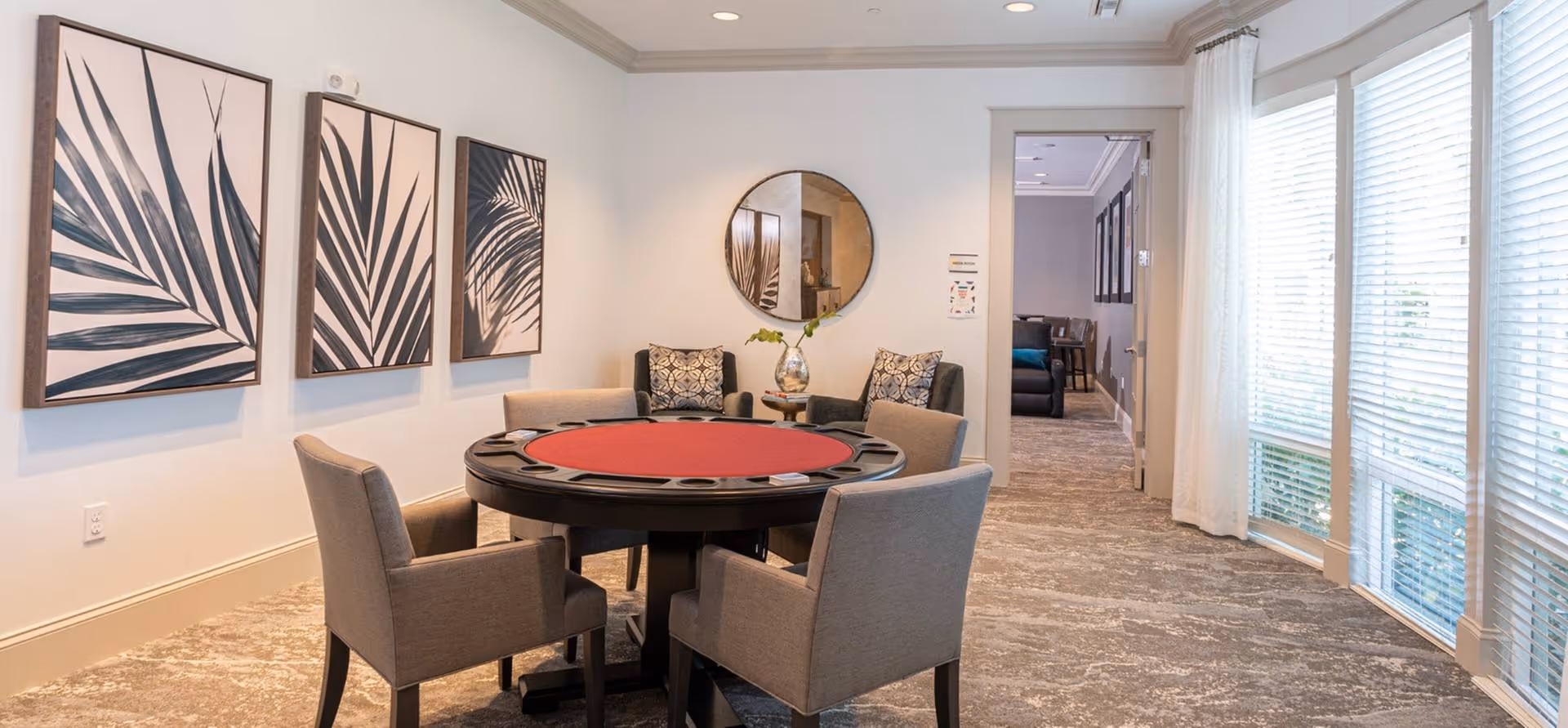 A well-lit interior room with a round poker table covered in red felt surrounded by four beige upholstered chairs. On the wall to the left, there are three framed artworks featuring abstract black and white leaf designs. A round mirror hangs on the far wall above two armchairs with patterned cushions and a small table with a decorative vase. Large windows with white blinds and sheer curtains line the right side of the room, allowing natural light to fill the space.