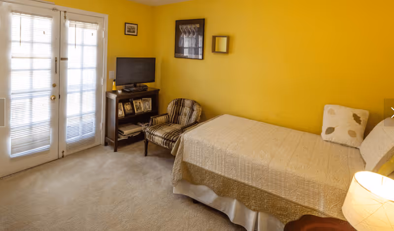A cozy bedroom with yellow walls featuring a single bed with a beige quilt and a pillow, a striped armchair, a small TV on a stand with framed photos, and French doors with blinds letting in natural light.