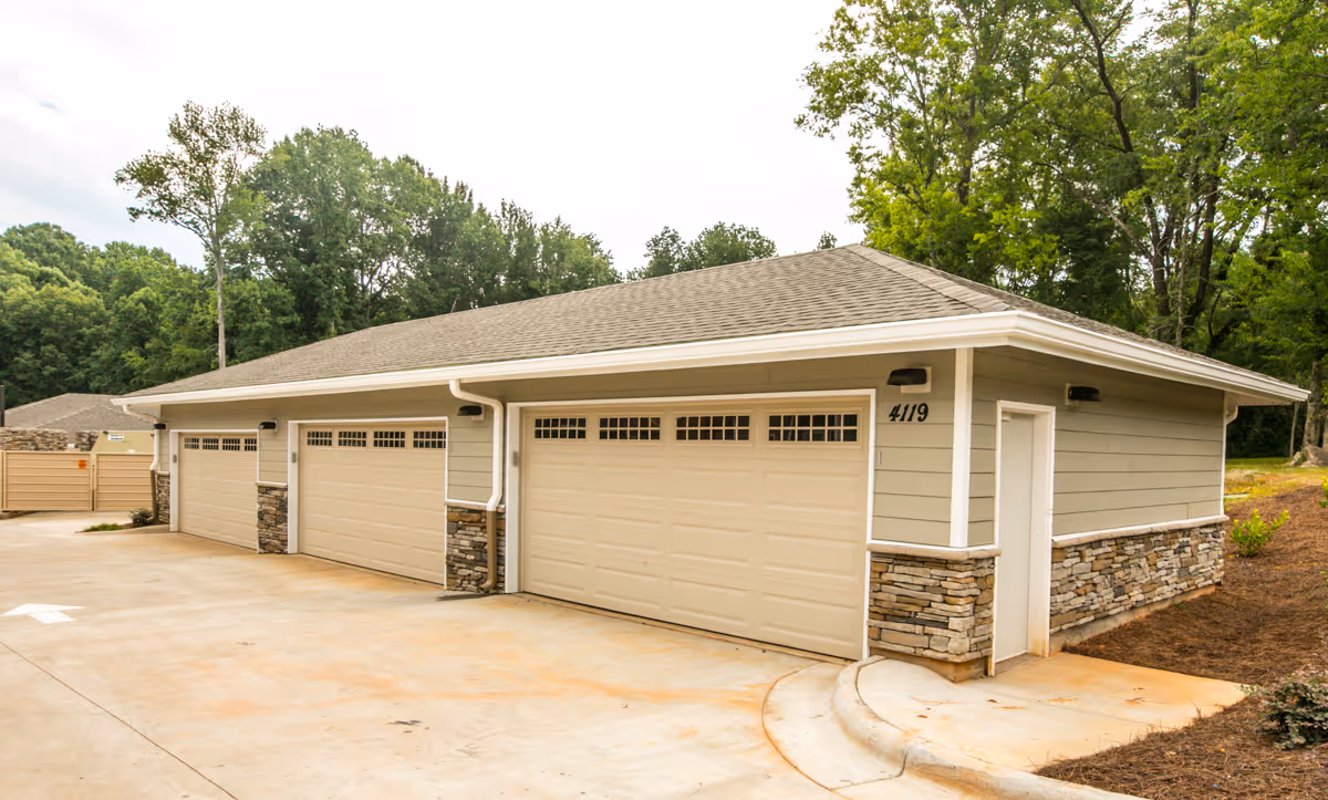 Exterior view of a building with four beige garage doors and stone accents on the lower walls, surrounded by trees and a concrete driveway.