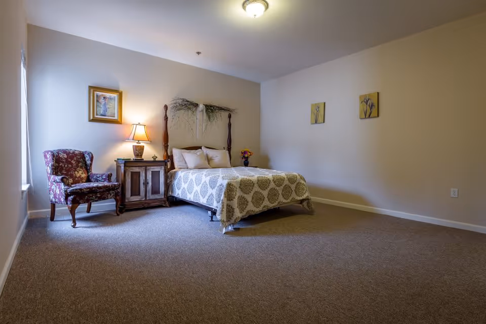 A bedroom with a bed covered in a patterned quilt, two pillows, and a decorative wall hanging above the headboard. To the left of the bed is a wooden nightstand with a lamp and some books, and next to it is a floral upholstered armchair. The room has beige walls, carpeted floor, and three framed pictures on the walls.