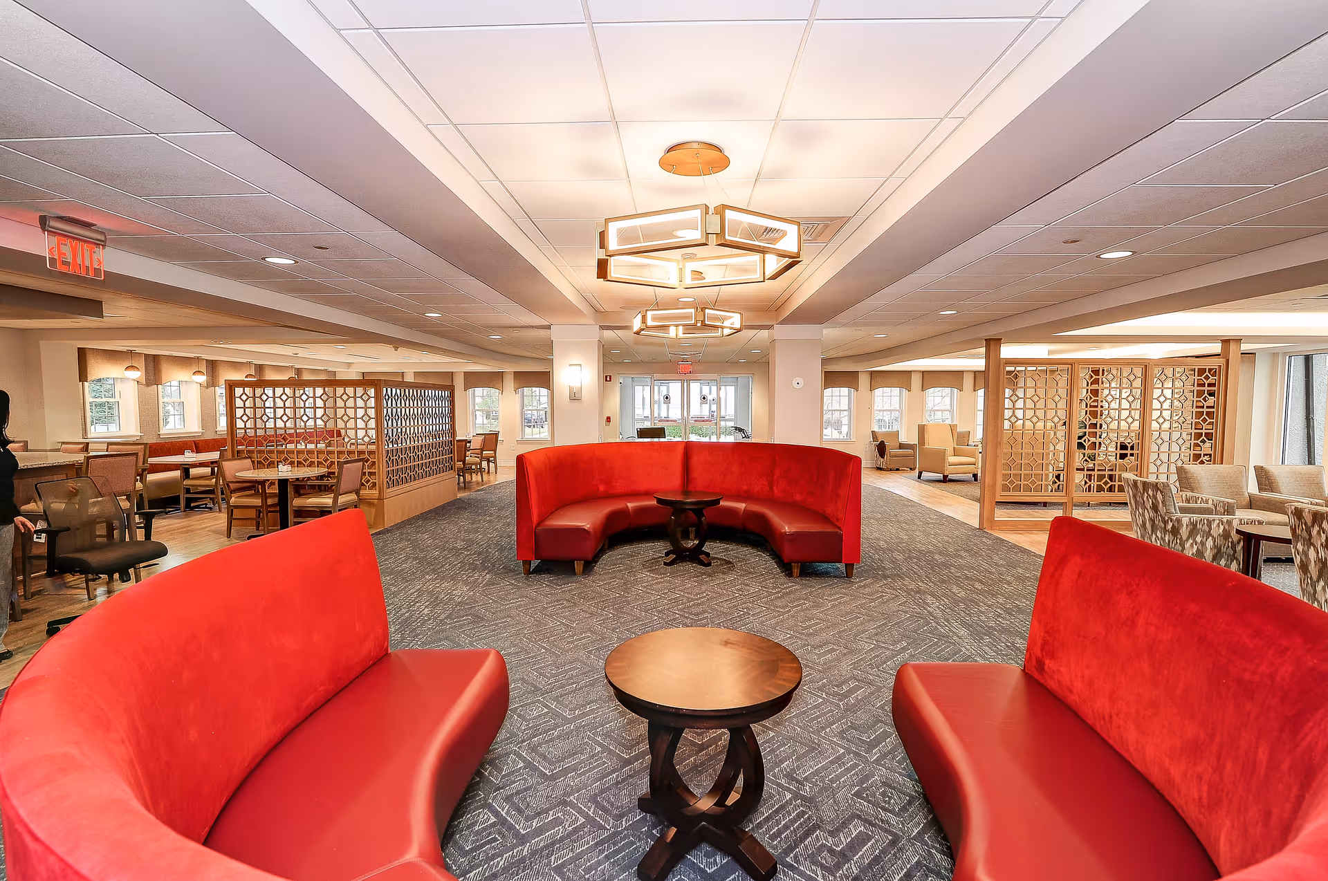Spacious and well-lit common area in a senior living facility featuring curved red upholstered seating around small wooden tables, with additional dining tables and chairs separated by decorative wooden partitions in the background. Modern ceiling lights and large windows provide ample lighting.