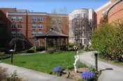 Outdoor courtyard area of a senior living facility with a green lawn, a wooden gazebo, paved walkways, flower beds with purple flowers, and surrounding multi-story brick buildings under a clear sky.