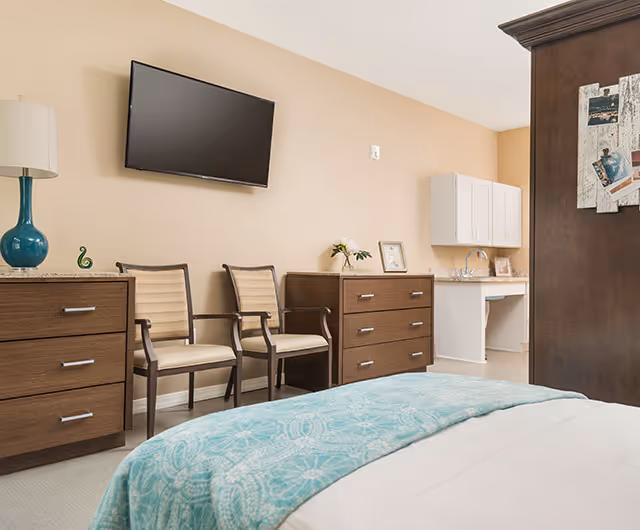 Bright bedroom with a bed topped by a teal patterned throw, a wall-mounted TV, two chairs, wooden dressers, and a small sink/cabinet area against a beige wall.