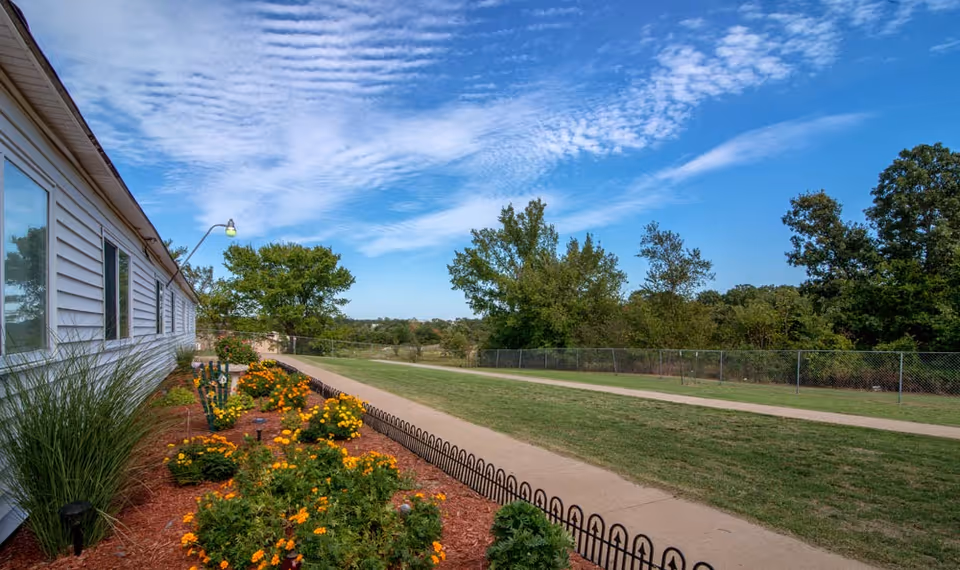 A landscaped outdoor area beside a white building with windows, featuring a flower bed with yellow and orange flowers, a paved walking path, green grass, and trees under a blue sky with scattered clouds.