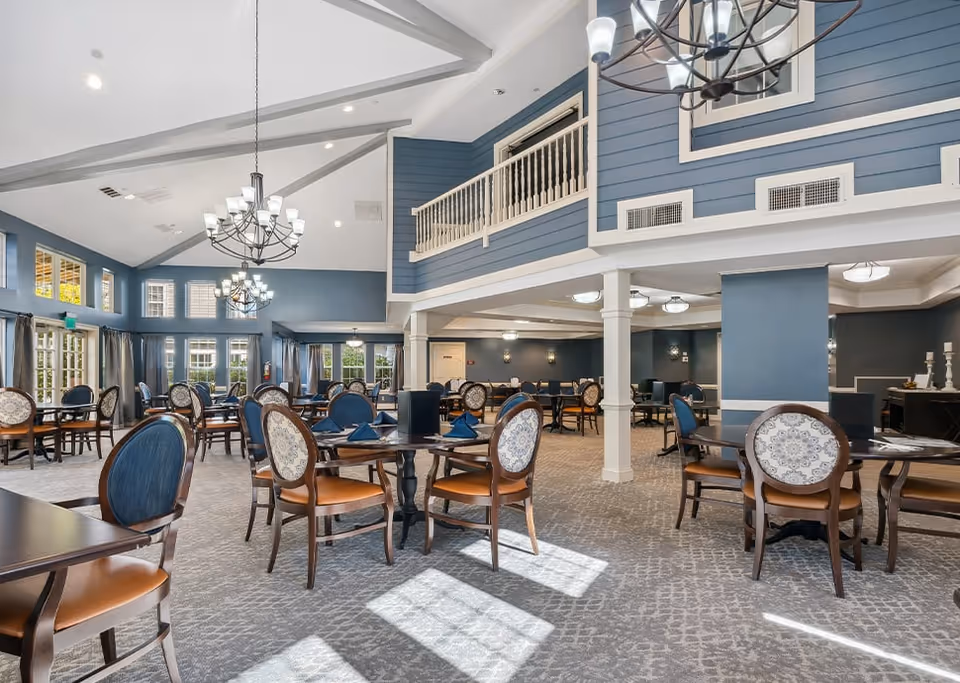 A spacious and well-lit dining room in a senior living facility with multiple round and rectangular tables surrounded by wooden chairs with blue and patterned upholstery. The room features high vaulted ceilings with exposed beams, large windows allowing natural light to fill the space, and elegant chandeliers hanging from the ceiling. The walls are painted in a soft blue color, and there is a balcony railing visible on the upper level.