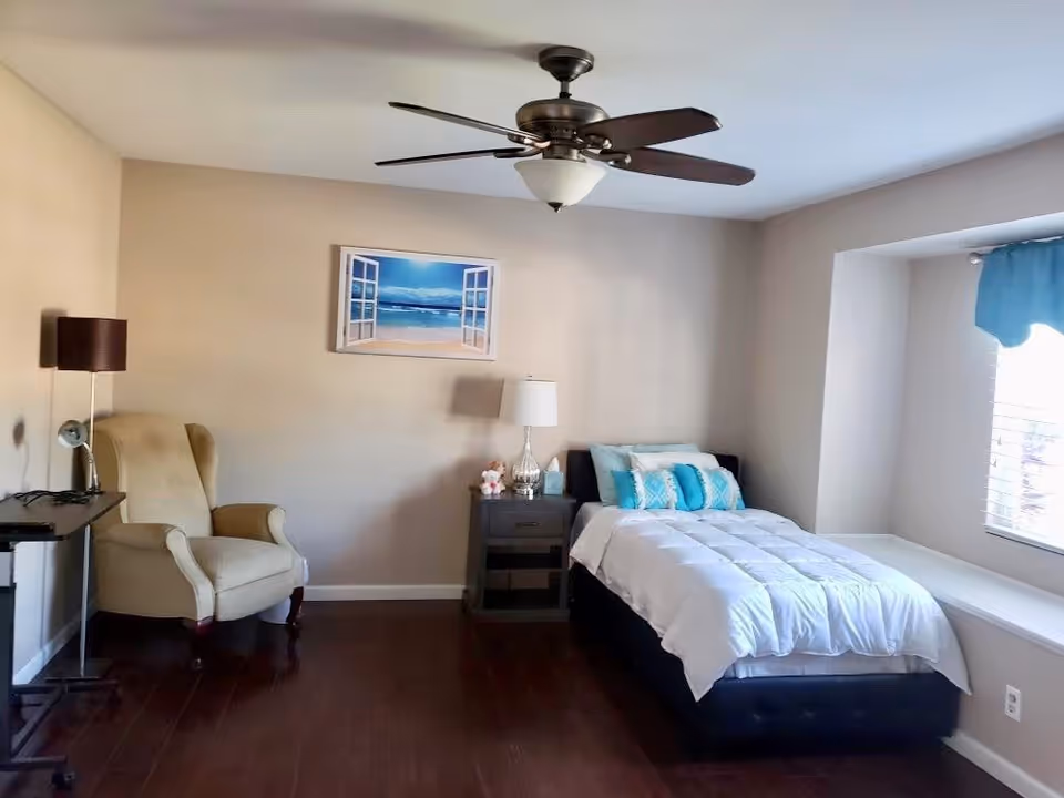 A cozy bedroom with a single bed covered in a white comforter and blue accent pillows. Next to the bed is a nightstand with a lamp and decorative items. There is a beige armchair and a small table with a lamp on the left side. A ceiling fan with lights is mounted on the ceiling, and a window with blue curtains lets in natural light.