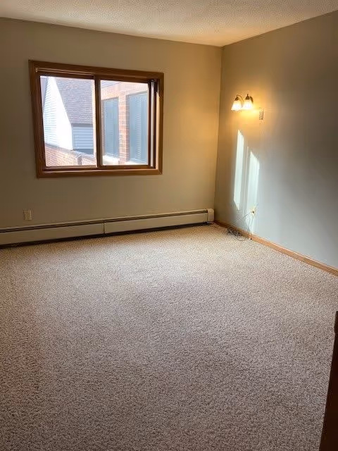 Empty carpeted room with a window, baseboard heater, and a wall sconce casting light on the pale walls.