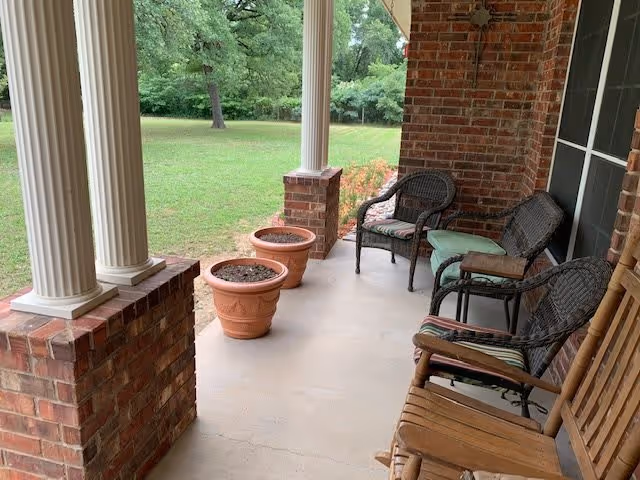 A covered porch area with brick pillars and a concrete floor. There are three empty terracotta plant pots lined up along the edge of the porch. Several chairs, including wicker chairs with cushions and a wooden rocking chair, are arranged along the porch next to a window. In the background, there is a grassy yard with trees.