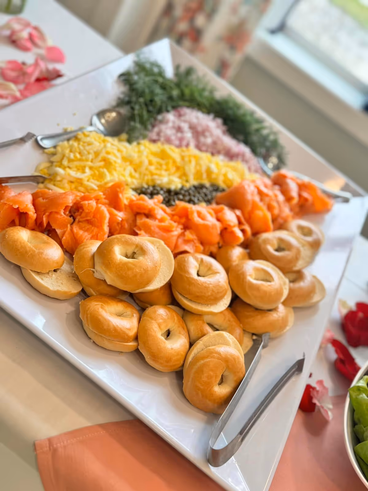 Platter of mini bagels with smoked salmon, shredded cheese, capers, chopped onion and herbs set on a buffet table.