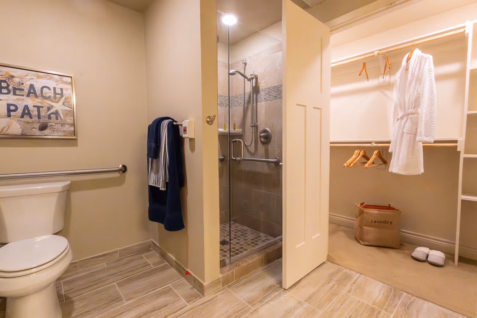 A bathroom with a toilet, a glass-enclosed shower with a showerhead and grab bars, and an adjacent walk-in closet containing a white robe, wooden hangers, a laundry basket, and a pair of slippers. The bathroom has beige walls, tiled floors, and a wall decoration that reads 'BEACH PATI'.