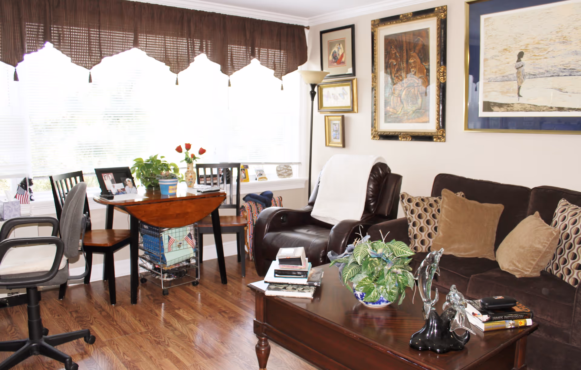 A cozy living room with a dark brown sofa adorned with patterned and solid pillows, a dark leather armchair with a white throw draped over it, and a wooden coffee table with books, a decorative sculpture, and a potted plant. There is a small wooden table with chairs near a large window covered with blinds and a brown valance. The walls are decorated with framed artwork and a floor lamp stands in the corner.