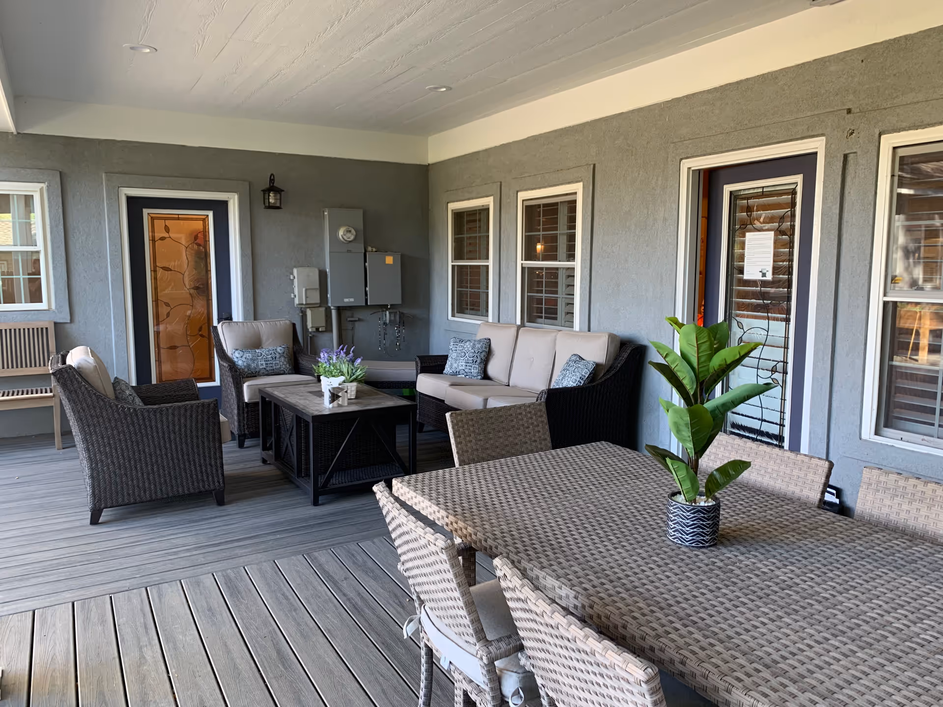 Covered outdoor patio area with wicker furniture including a sofa, armchairs, and a coffee table with a small plant. There is also a dining table with chairs and a potted plant on top. The area has gray walls with windows and doors, and a wooden floor.