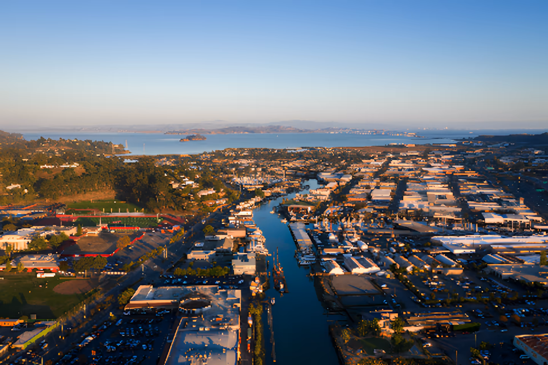 Aerial view of a coastal city with a waterway running through the center, surrounded by buildings, greenery, and distant hills under a clear blue sky.