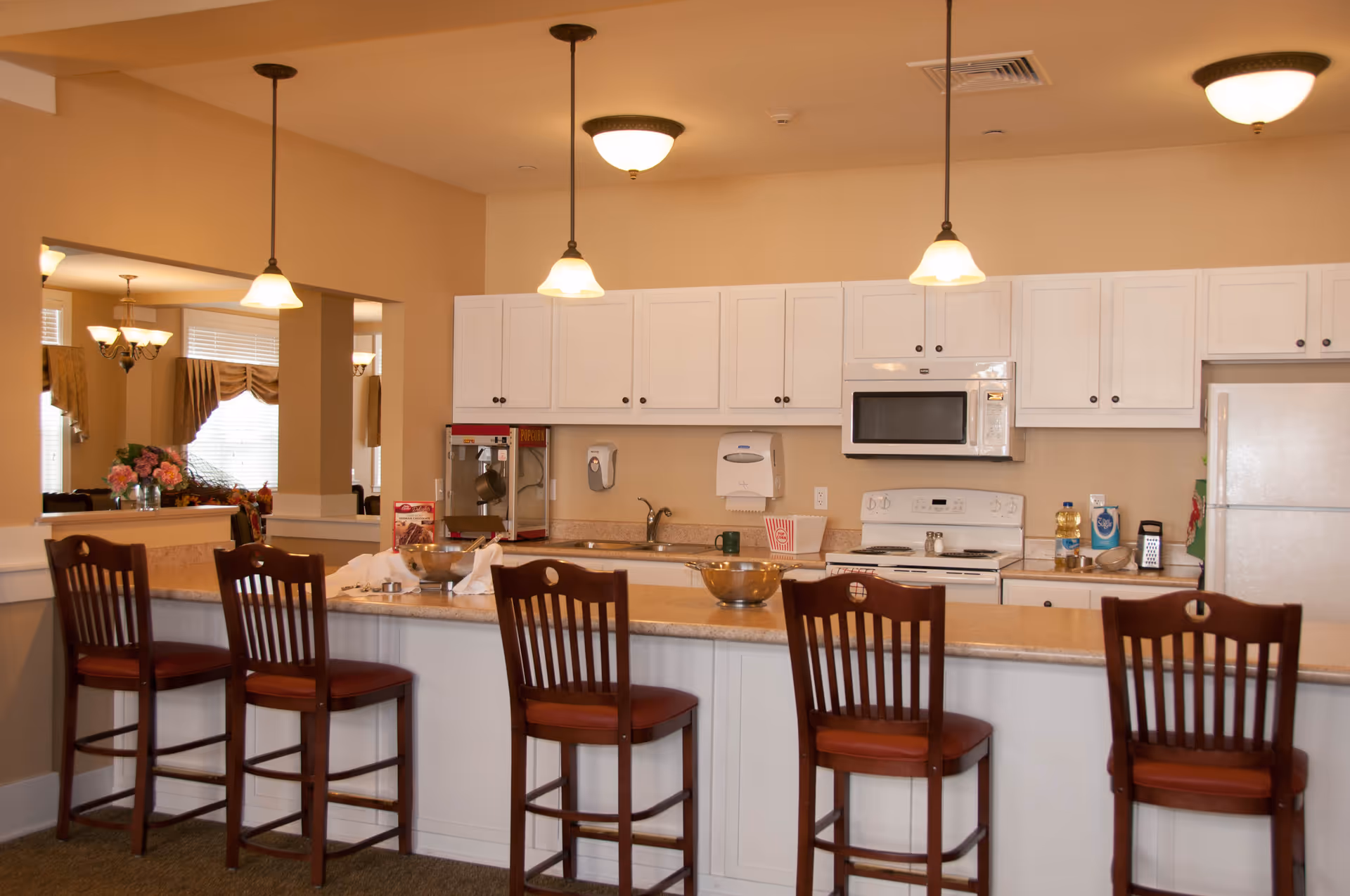 A kitchen area with a long counter and five wooden bar stools with red cushions. The kitchen has white cabinets, a white refrigerator, a white microwave, and a white stove. There are three pendant lights hanging above the counter and a popcorn machine on the counter near the sink. In the background, there is a dining area with a chandelier and windows with curtains.