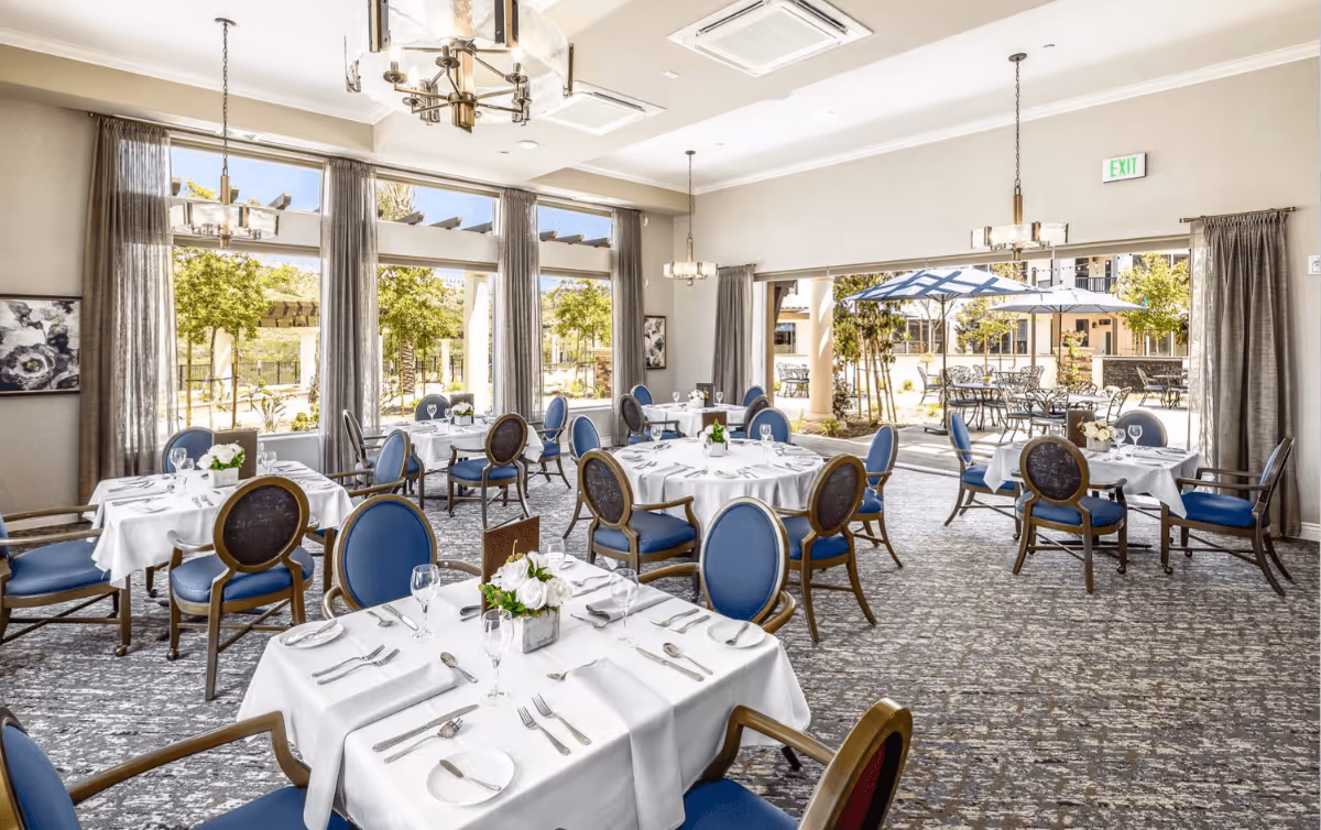 A bright and elegant dining room with round and square tables covered with white tablecloths, set with silverware, plates, and glasses. Blue cushioned chairs surround the tables. Large windows and glass doors provide a view of an outdoor patio area with tables, chairs, and umbrellas. The room features modern chandeliers and neutral-colored walls with curtains.