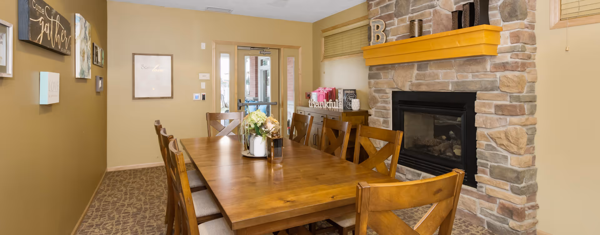A cozy dining room with a long wooden table surrounded by wooden chairs. On the table is a decorative centerpiece with flowers. The room features a stone fireplace with a wooden mantel decorated with various items including a letter B. The walls are painted a warm beige color and adorned with framed artwork and signs. A glass door at the far end leads outside.