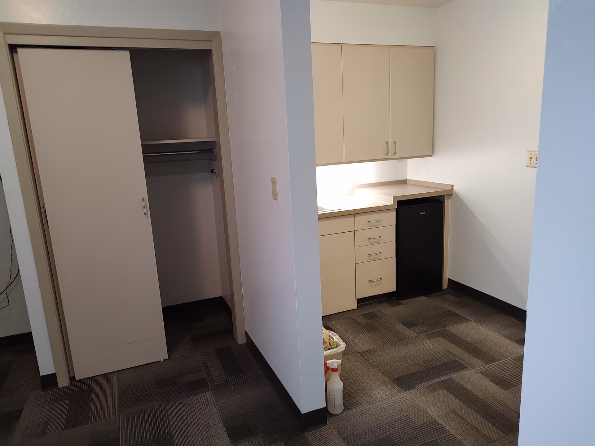 Carpeted assisted living area showing a small kitchenette with beige cabinets, a mini fridge and drawers beside an open sliding-door closet.