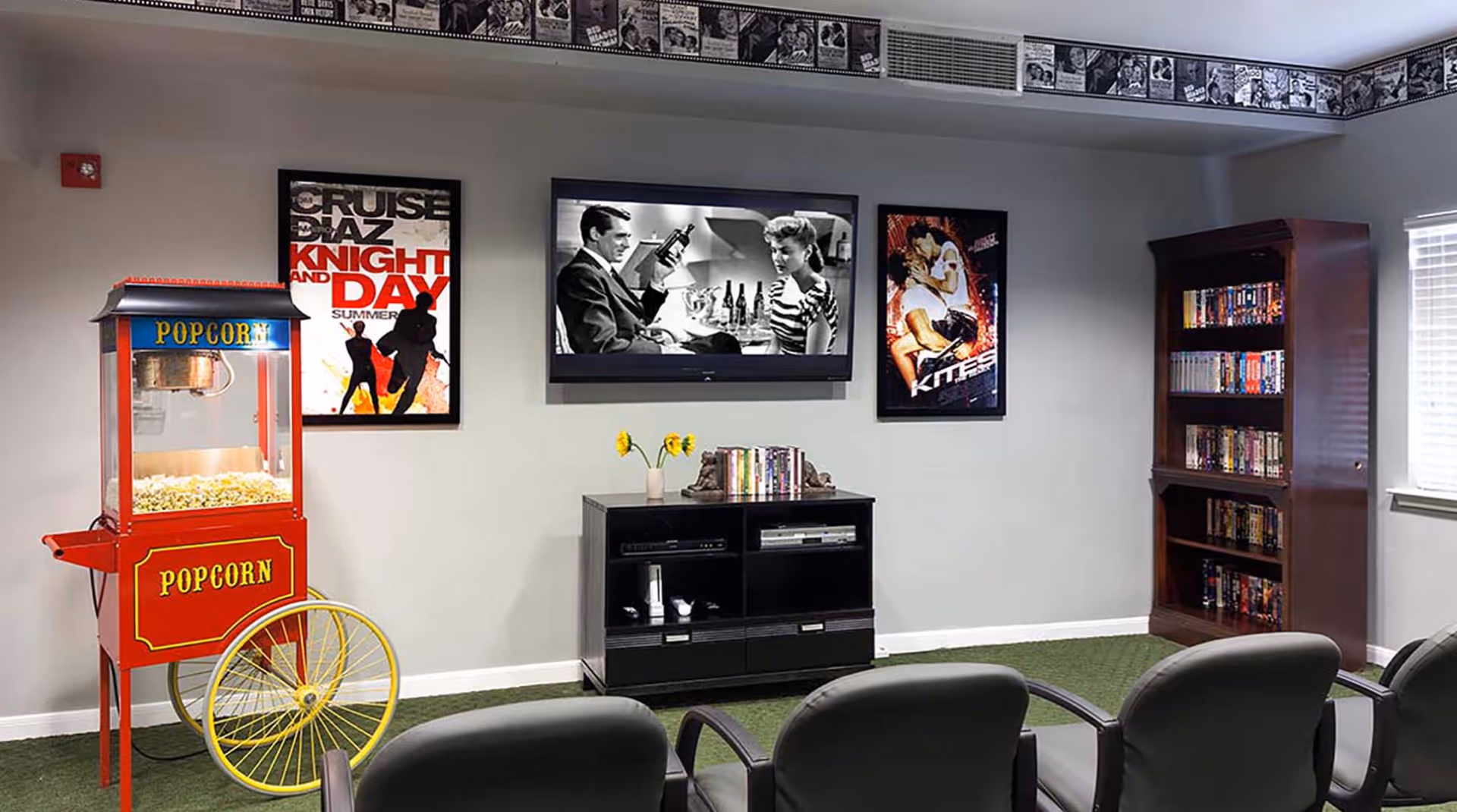 A small movie theater room with a popcorn machine on the left, a flat-screen TV mounted on the wall showing a black and white movie, movie posters on the wall, a black media cabinet with DVDs and a vase with yellow flowers, a bookshelf filled with DVDs, and several black chairs facing the TV.