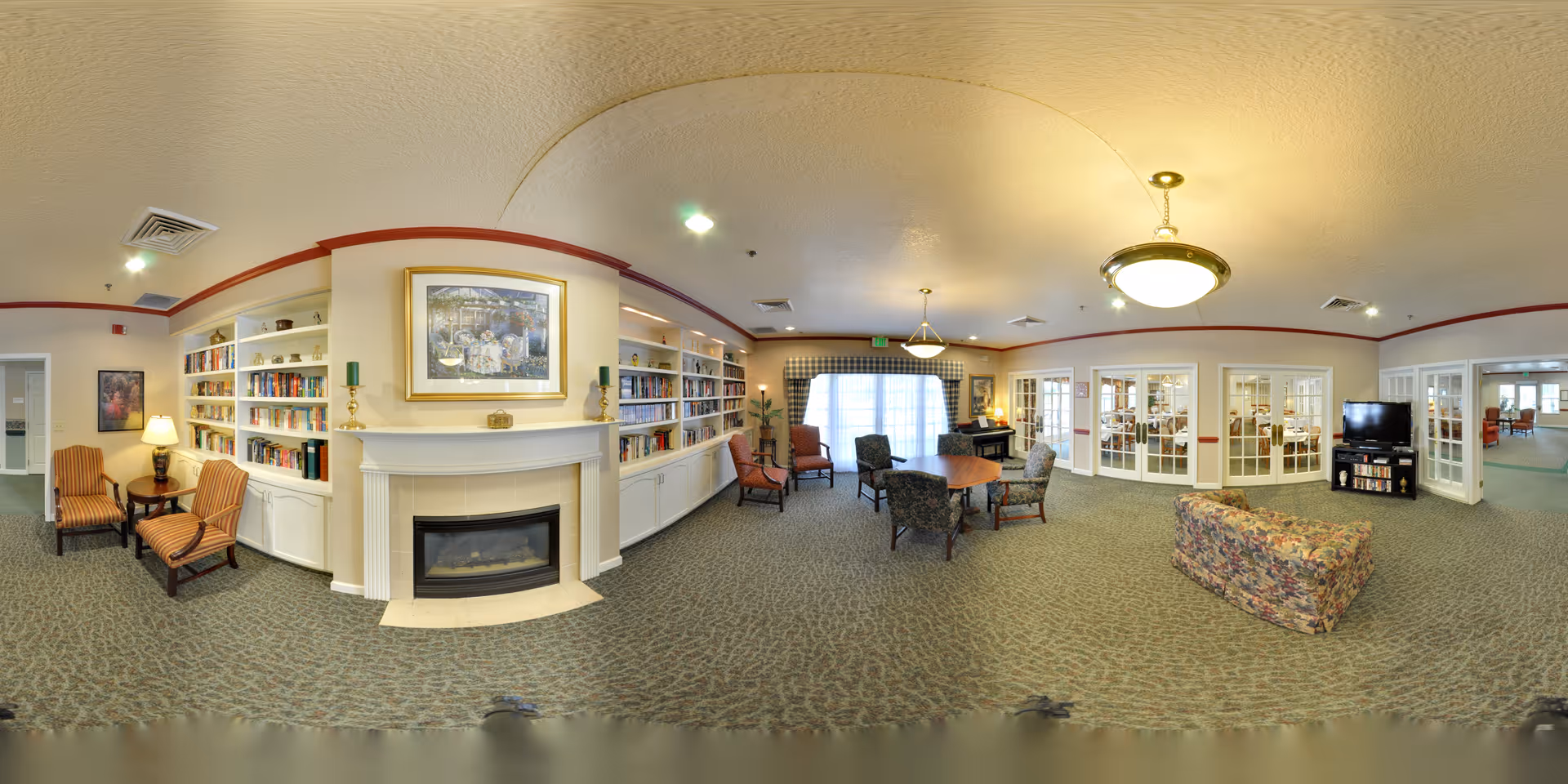 A spacious senior living facility common area with built-in bookshelves filled with books, a white fireplace with a framed painting above it, several armchairs and a floral-patterned sofa, a round table with chairs, a piano near large windows with curtains, and a television on a stand. The room has carpeted floors, soft lighting, and glass-paneled doors leading to other rooms.