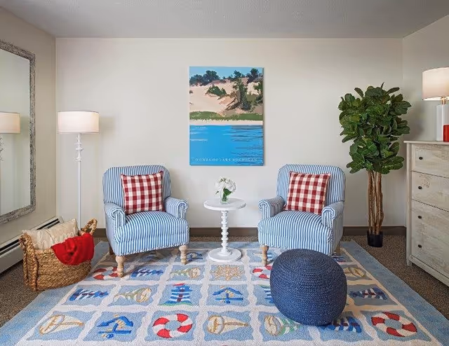 A cozy sitting area with two blue and white striped armchairs each with a red and white checkered pillow. Between the chairs is a small white round table with a flower vase. A large blue knitted pouf is in front of the chairs on a colorful rug with nautical designs. To the left is a tall white floor lamp and a basket with blankets. On the right is a tall green plant and a white wooden dresser with a lamp on top. A painting of a lakeside scene hangs on the wall behind the chairs.
