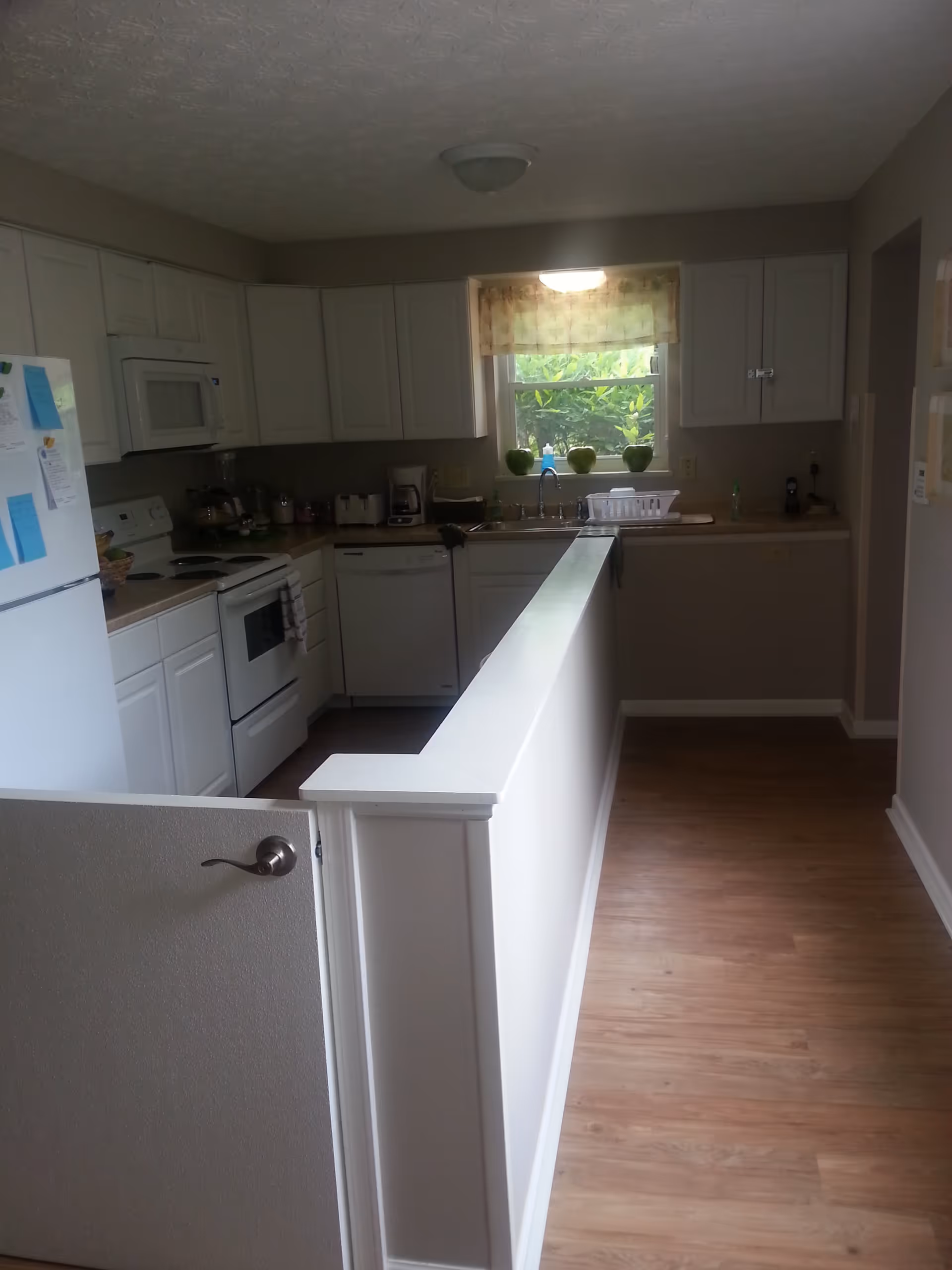 A clean kitchen with white cabinets, a white refrigerator, stove, microwave, dishwasher, and a sink under a window with a floral curtain. The kitchen has a half wall with a gate and wood flooring.