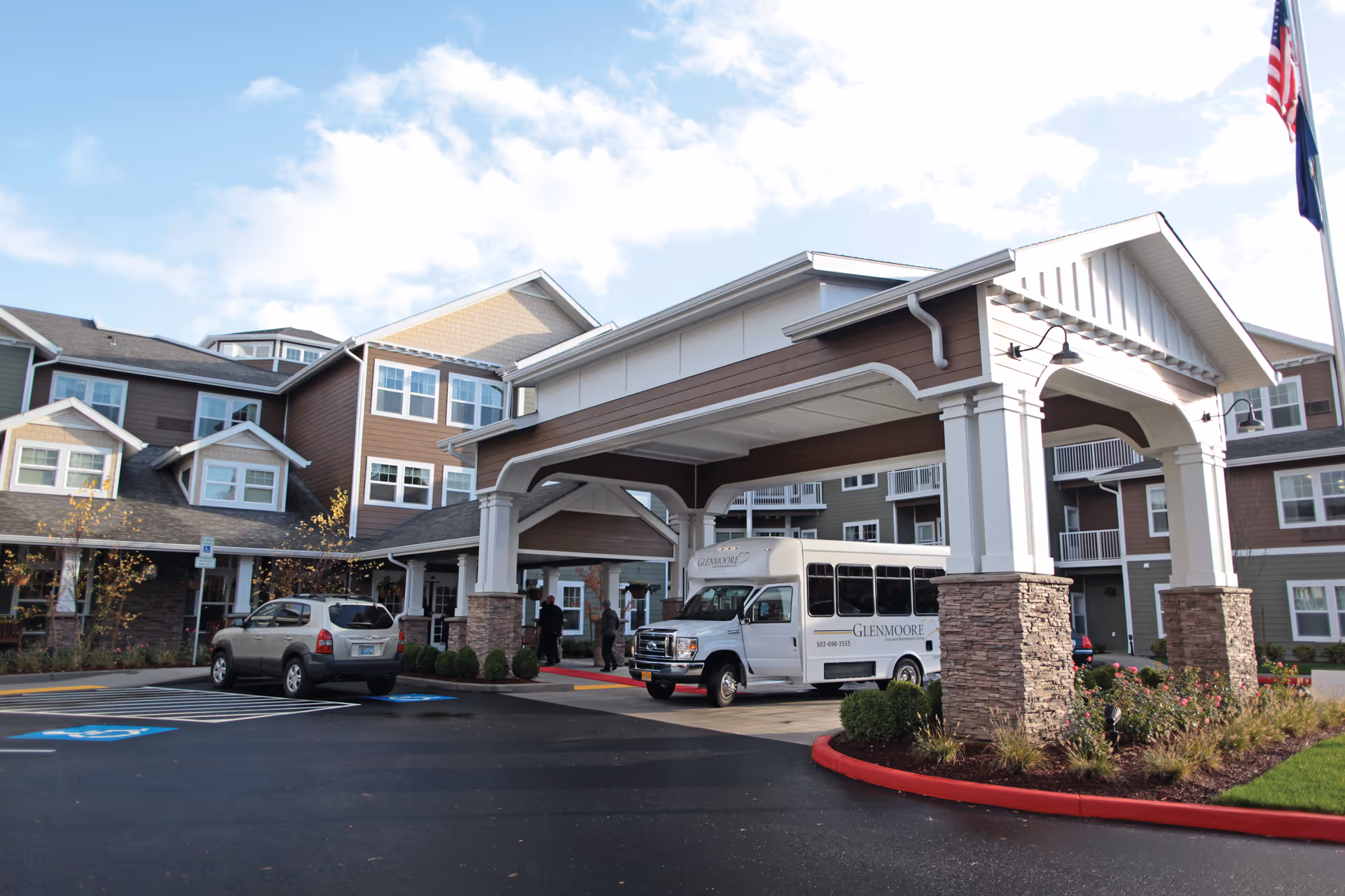 Exterior view of Glenmoore Gracious Retirement Living facility showing a covered entrance with stone pillars, a white shuttle bus parked under the entrance canopy, a silver SUV in the parking lot, and a multi-story building with brown and beige siding in the background under a partly cloudy sky.