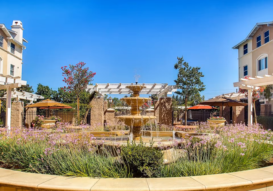 A multi-tiered fountain in a sunny landscaped courtyard with flowering plants, umbrellas, and surrounding apartment buildings.