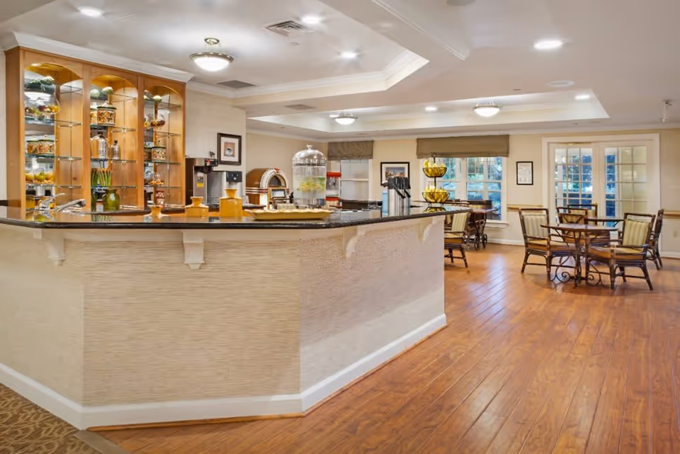 A bright and spacious dining area in a senior living facility featuring a large counter with a black countertop, glass shelves with decorative items, a water dispenser, and several wooden tables with chairs arranged around the room. The floor is wooden, and the walls are light-colored with windows and framed pictures.