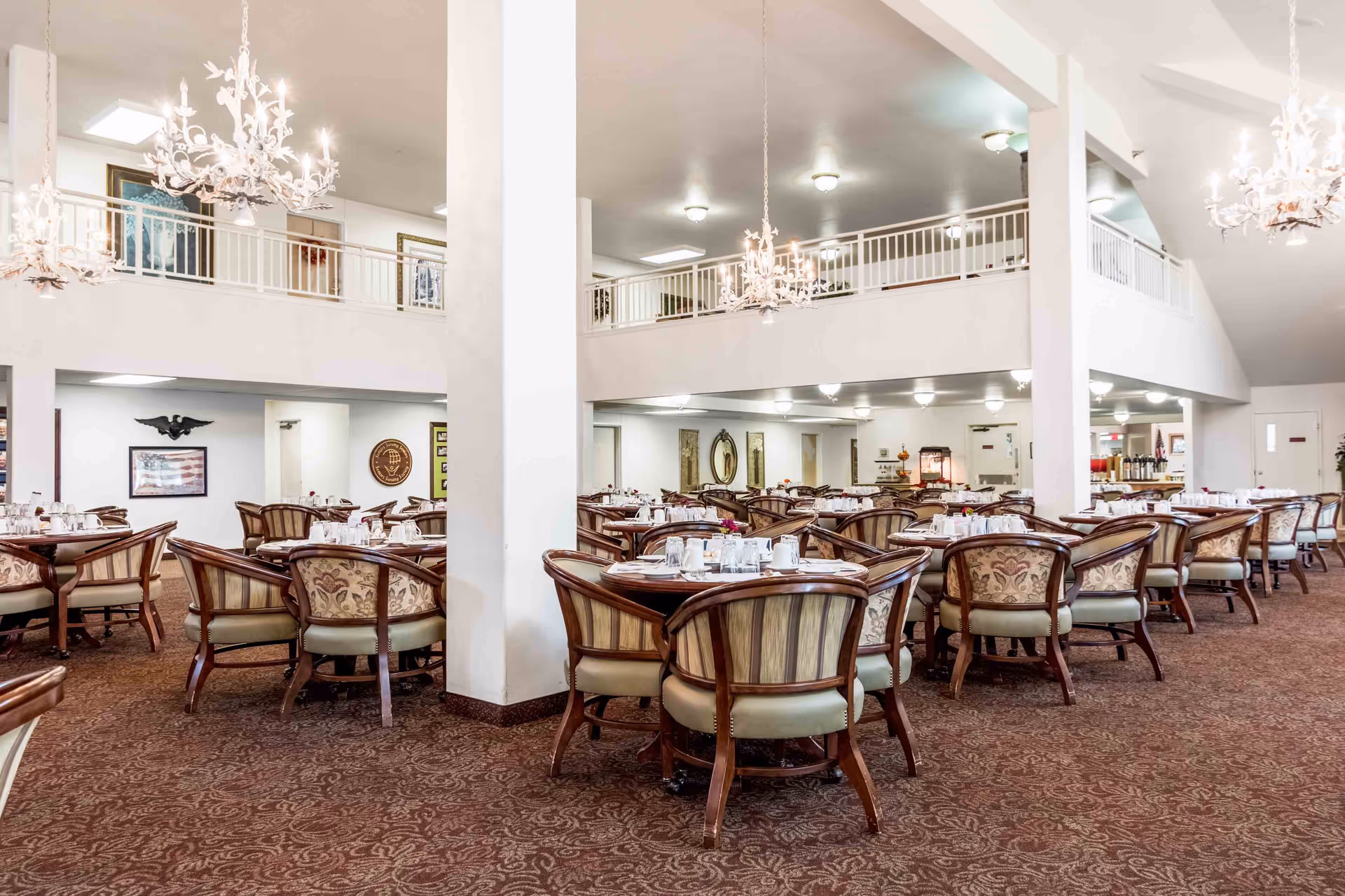 A spacious dining room with multiple round tables surrounded by cushioned wooden chairs. The room features a high ceiling with white chandeliers, a patterned carpet, and a second-floor balcony overlooking the dining area. The tables are set with cups, glasses, and napkins, ready for a meal.