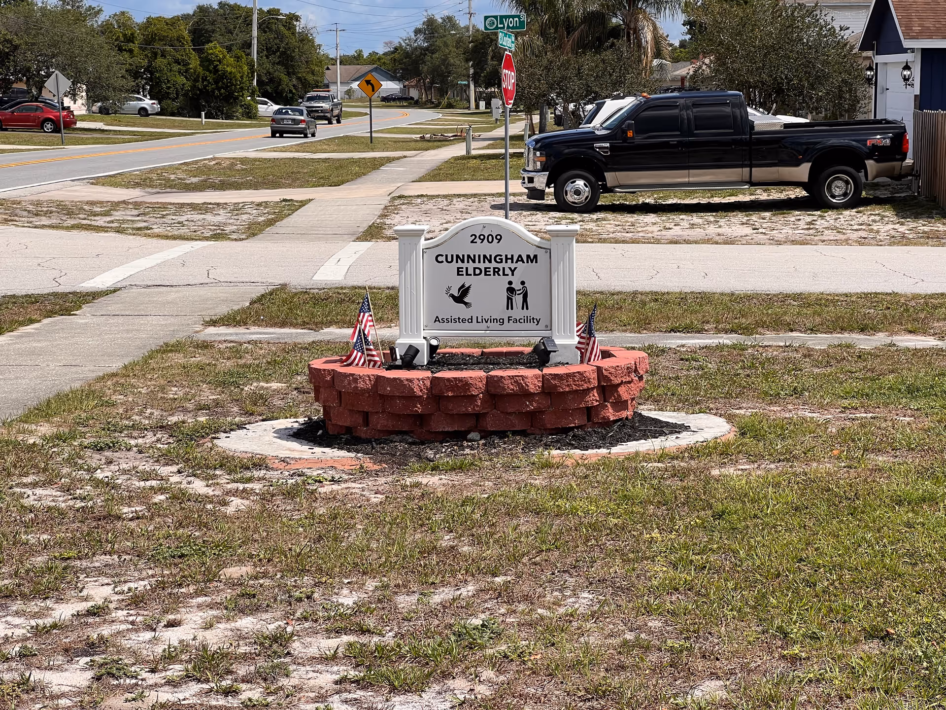 Outdoor view of a street corner with a sign for Cunningham Elderly Assisted Living Facility placed on a circular red brick base with small American flags. There are sidewalks, a stop sign, street signs, parked vehicles, and houses in the background under a partly cloudy sky.