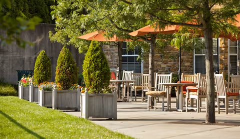 Sunlit outdoor patio with wooden tables and chairs, orange umbrellas, and planter boxes in front of a stone building.