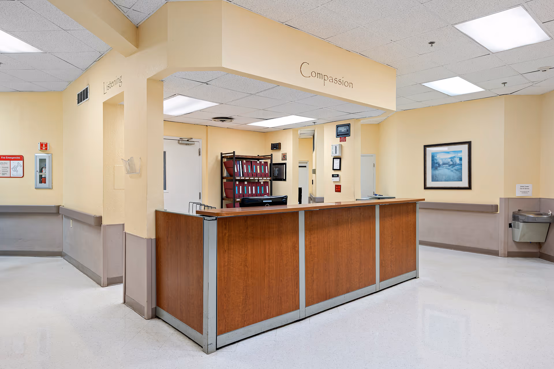 Reception desk area in a senior living facility with beige walls, a wooden counter, and fluorescent ceiling lights. The words 'Listening' and 'Compassion' are written on the walls above the desk. There is a fire emergency box on the left wall, a water fountain on the right, and a framed picture on the far wall.