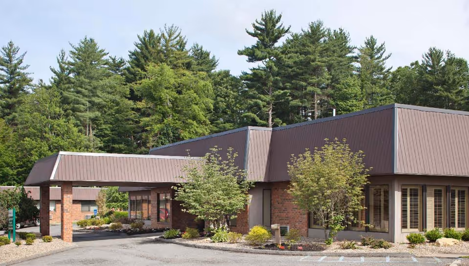 Exterior view of a single-story brick building with a brown sloped roof, surrounded by landscaped greenery and trees in the background. The building has large windows with blinds and a covered entrance driveway.
