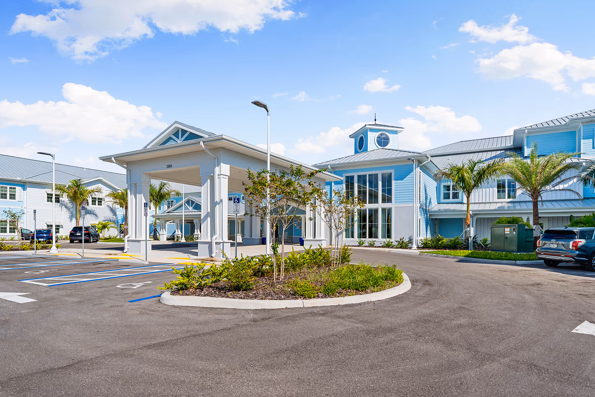 Exterior view of The Meridian at Punta Gorda Isles senior living facility showing a light blue building with white trim, a covered entrance, palm trees, and a parking lot with several cars under a partly cloudy sky.