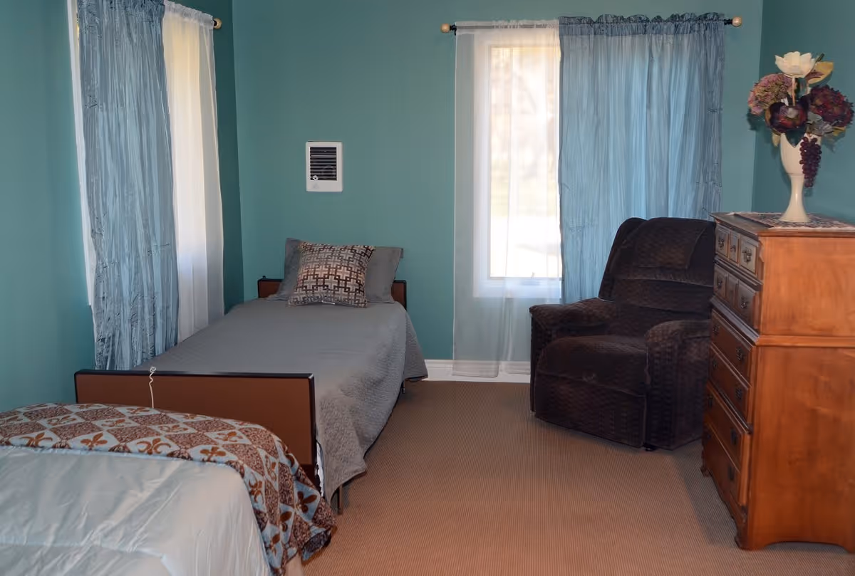 A bedroom with teal walls featuring two single beds, one with a gray bedspread and decorative pillows, and the other partially visible with a patterned blanket. There is a dark brown armchair near a window with sheer and blue curtains. A wooden dresser with a vase of flowers on top is also present.