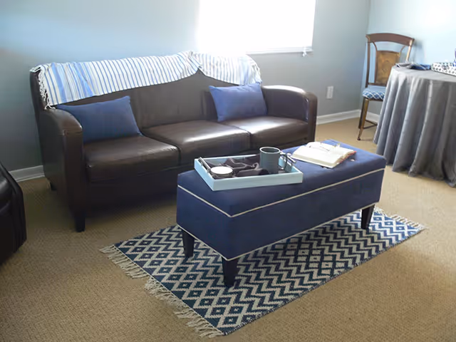 A cozy living room with a dark brown leather sofa adorned with two blue pillows and a striped throw blanket. In front of the sofa is a blue upholstered ottoman with a tray holding a cup and other items, placed on a patterned blue and white rug. To the right, there is a wooden chair and a round table covered with a gray tablecloth. The room has beige carpet and light blue walls with a window letting in natural light.