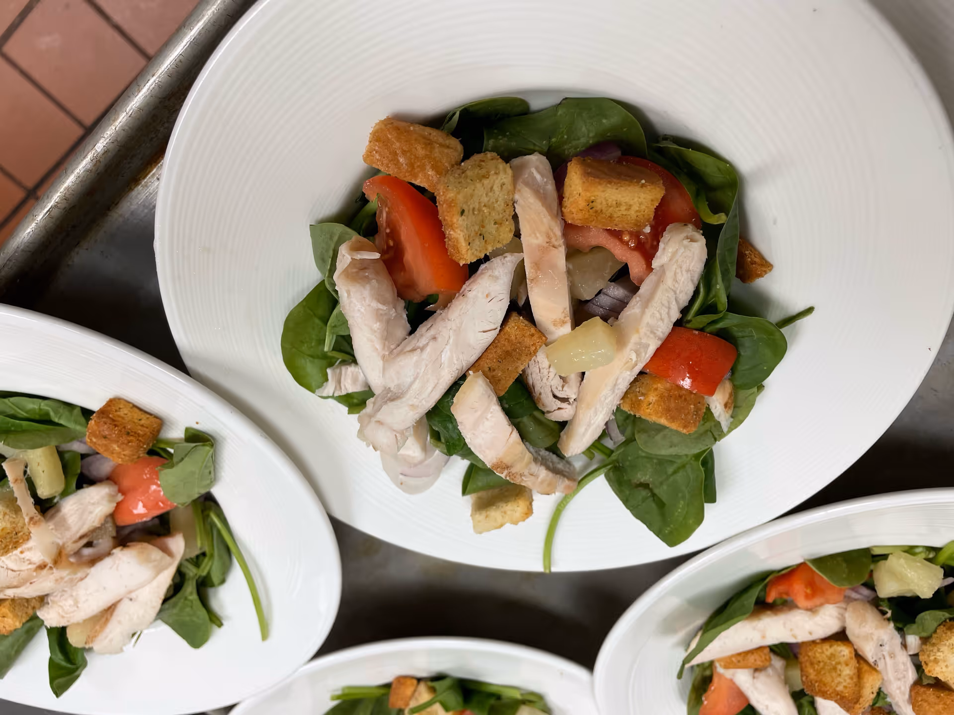 Close-up of several white plates each containing a fresh salad with spinach leaves, sliced grilled chicken, tomato wedges, croutons, pineapple chunks, and red onion, arranged on a metal surface.