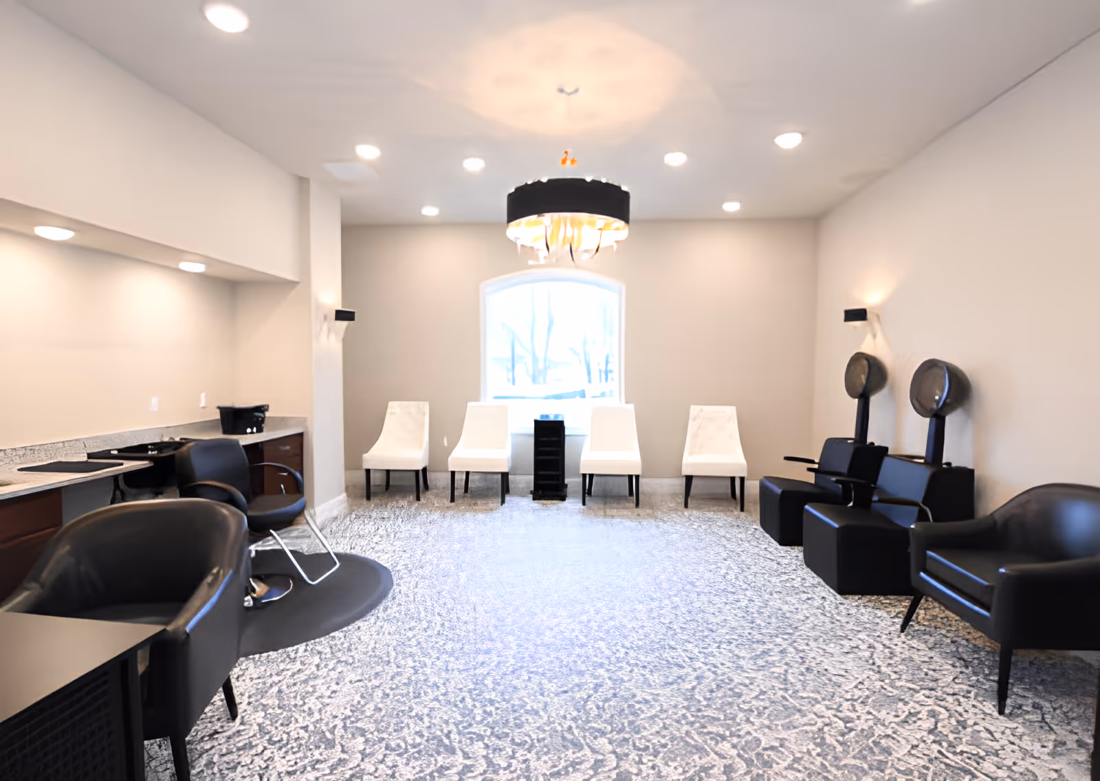 A bright and spacious salon room with a patterned carpet floor, four white chairs lined up against the back wall under a large window, two black hair dryer chairs on the right, and black salon chairs with counters on the left. A modern chandelier hangs from the ceiling in the center of the room.