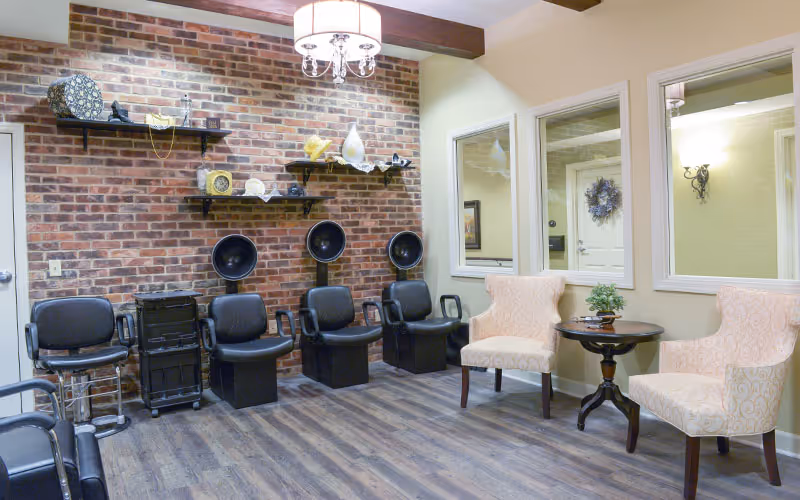 Interior of a salon area with three black hair dryer chairs against a brick wall, two beige patterned armchairs with a small round wooden table and a potted plant between them, wooden flooring, and large windows on the right wall reflecting the room.