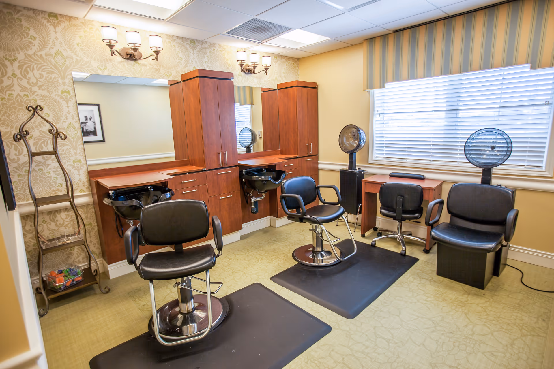 Interior view of a hair salon area in a senior living facility with two black salon chairs in front of wooden cabinets and sinks, two hair dryers on stands near a window with blinds, and a small desk with a chair.