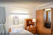 A small, neatly arranged bedroom with a single bed featuring white and blue patterned pillows. Next to the bed is a wooden nightstand with a lamp, framed photo, and a small decorative item. Above the bed hangs a framed picture of a purple flower. There is a wooden wardrobe with a mirror and a small television inside. The room has light-colored walls and a window with gray curtains.