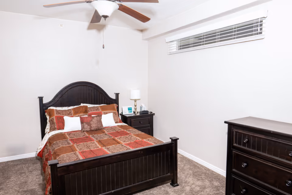 A bedroom with a dark wooden bed frame and a colorful patchwork quilt in shades of red, brown, and beige. The bed has multiple pillows, and there is a matching dark wooden nightstand with a lamp and some items on it. A dark wooden dresser is positioned against the right wall. The room has light-colored walls, a carpeted floor, a ceiling fan with wooden blades, and a narrow window with white blinds near the ceiling.