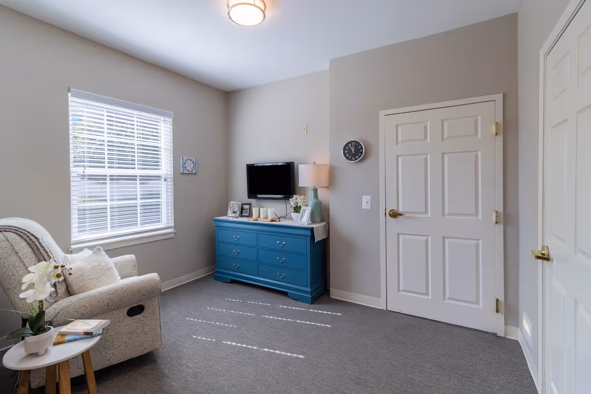 A cozy living room with a patterned armchair next to a small round side table holding a potted orchid and a book. A large window with white blinds lets in natural light. Across from the chair is a blue dresser with a flat-screen TV mounted above it, a table lamp, framed photos, and decorative candles. The walls are painted light gray, and there are two white doors with gold handles. A round wall clock is mounted on the wall near one of the doors.