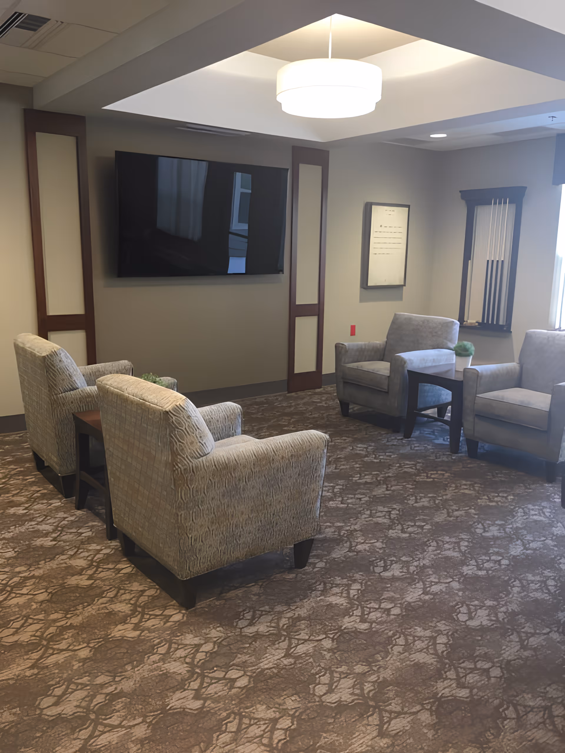A cozy seating area with four upholstered armchairs arranged around two small wooden side tables. A large flat-screen TV is mounted on the wall, and a modern ceiling light fixture hangs above. The room has carpeted flooring with a patterned design, neutral-colored walls, and a window letting in natural light. There is a wall-mounted rack holding pool cues in the corner.