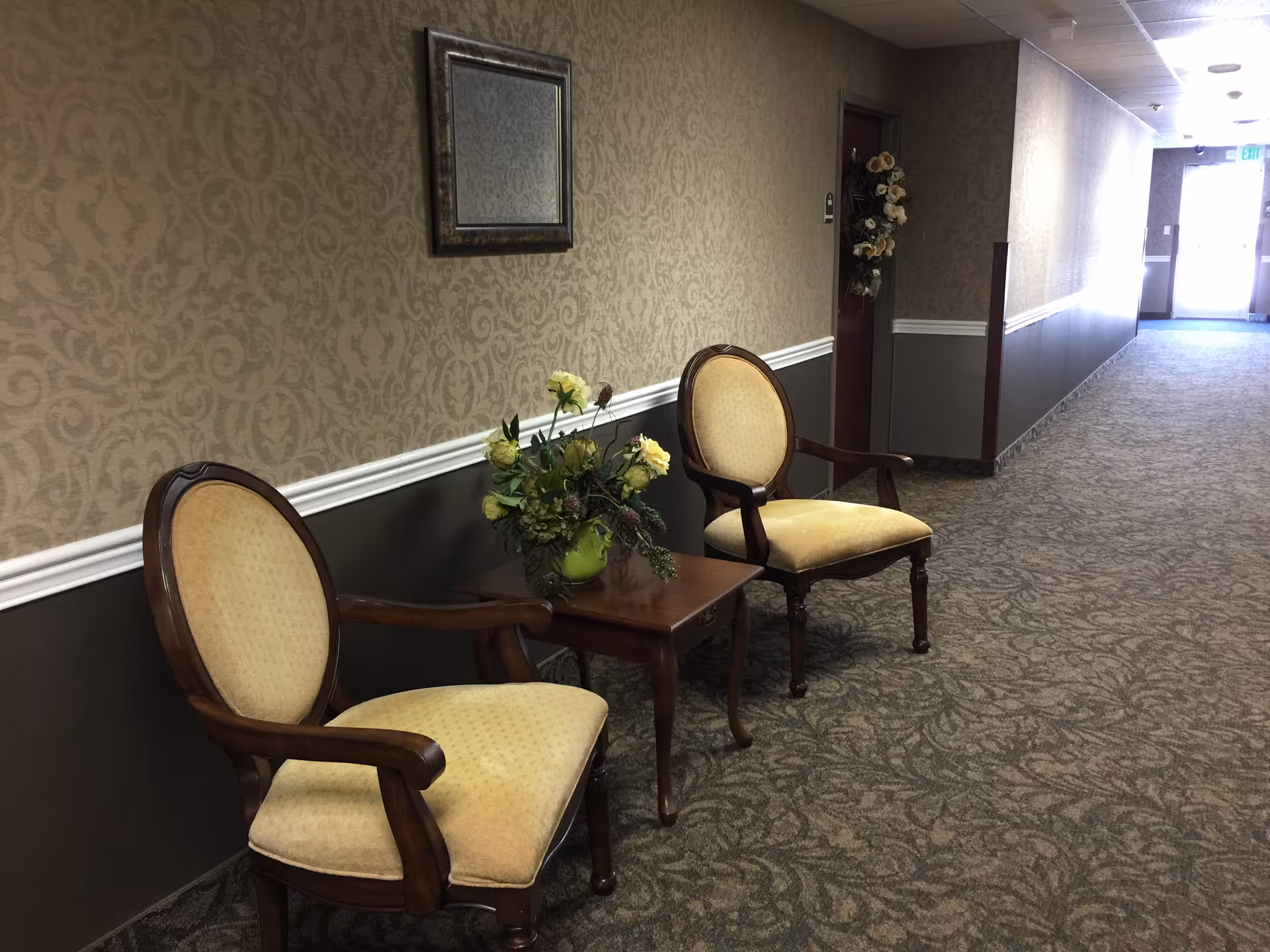 Two upholstered chairs and a small side table with a floral arrangement sit along a carpeted, wallpapered hallway.