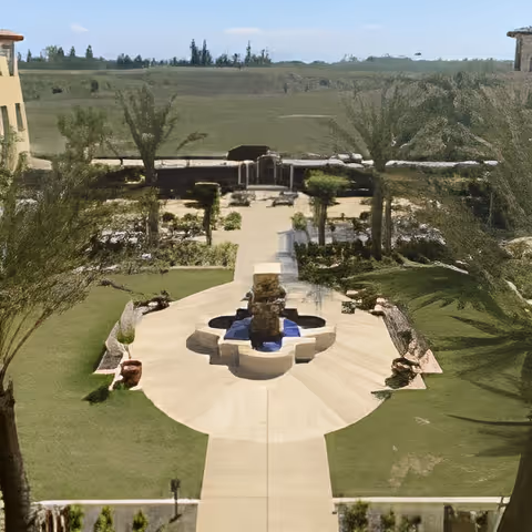 View of a landscaped outdoor garden area with a central stone fountain surrounded by pathways and greenery, with buildings on either side and a clear sky in the background.