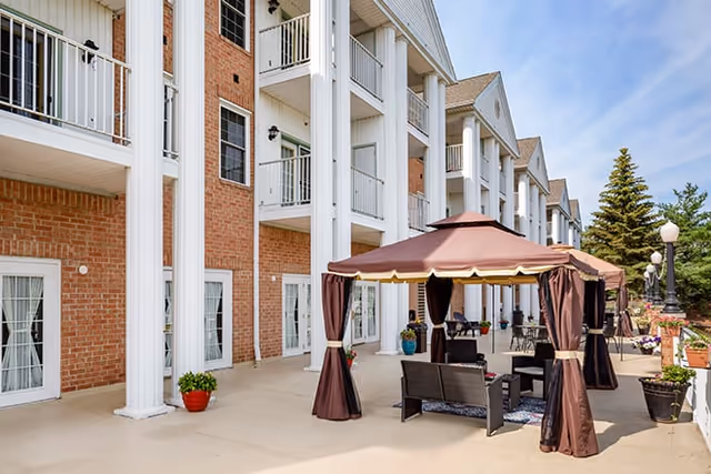 Outdoor patio area at Brookdale Camelot Medina featuring a brown canopy with curtains, outdoor seating including chairs and a table, potted plants, and a large brick building with white columns and balconies in the background under a clear blue sky.