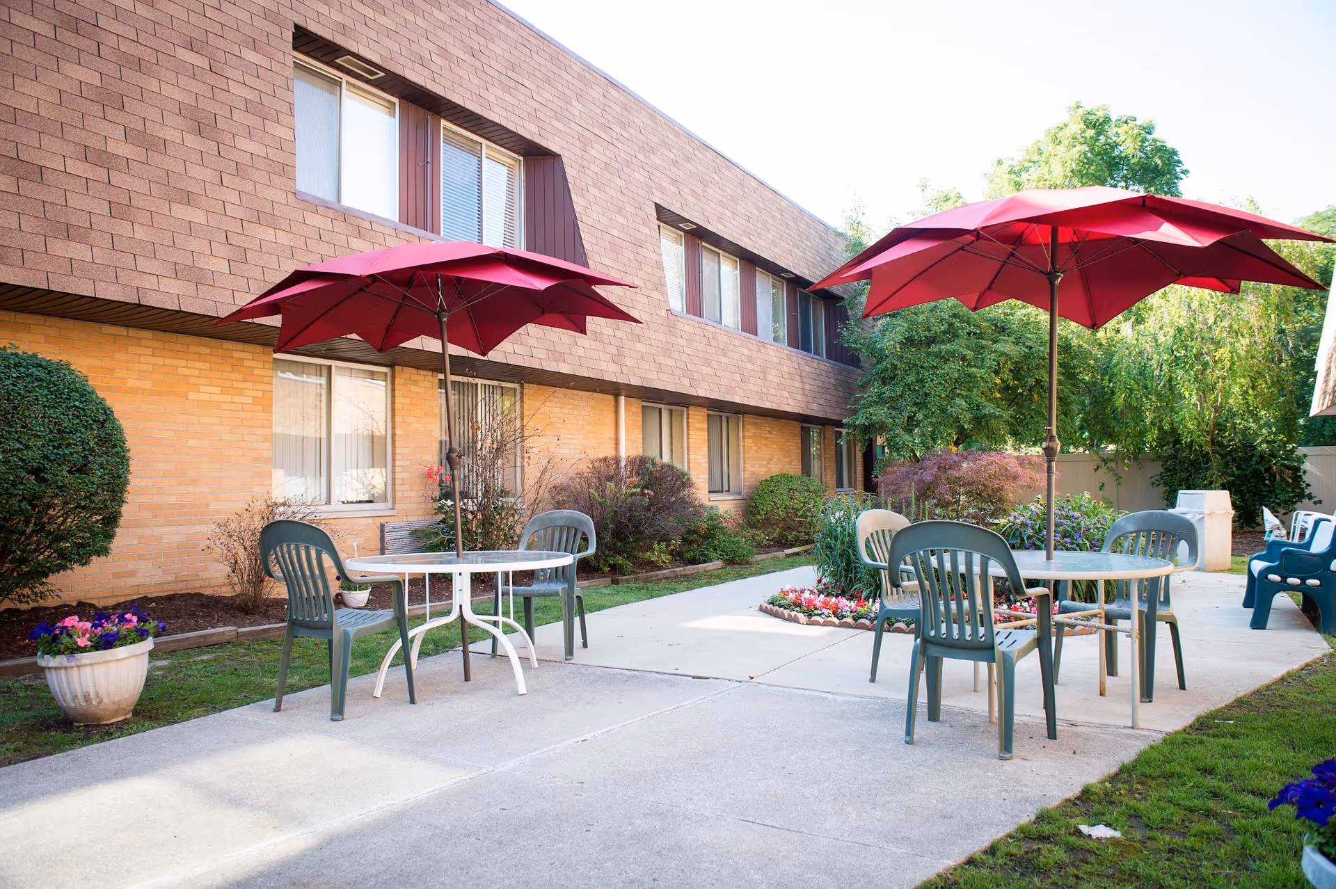 Outdoor patio area at Medilodge of Farmington with two round tables, each shaded by a red umbrella, surrounded by green plastic chairs. The patio is adjacent to a two-story brick building with windows, and there are various bushes, flowers, and trees around the paved walkway.