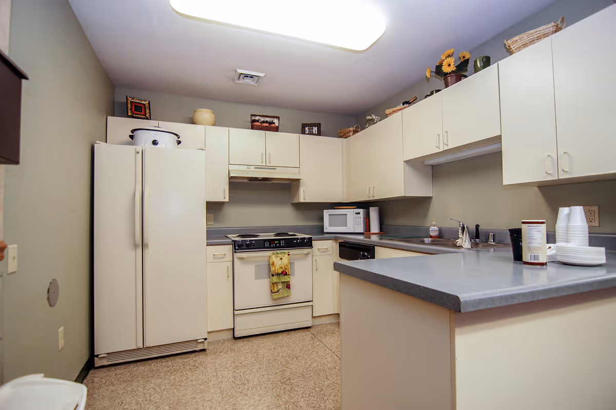 A clean kitchen with white cabinets and gray countertops. The kitchen includes a white refrigerator, a stove with an oven, a microwave, a dishwasher, and a sink. There are decorative items on top of the cabinets and some kitchen essentials like paper cups and plates on the counter.