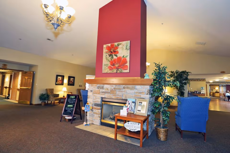 A cozy assisted living facility common area with a central stone fireplace topped by a red wall section and a painting of red flowers. There is a small wooden table with framed pictures and a 'Home Sweet Home' sign, a potted plant, and a blue upholstered chair nearby. In the background, there is a hallway, a chalkboard menu, and additional seating areas with lamps and framed artwork on the walls.